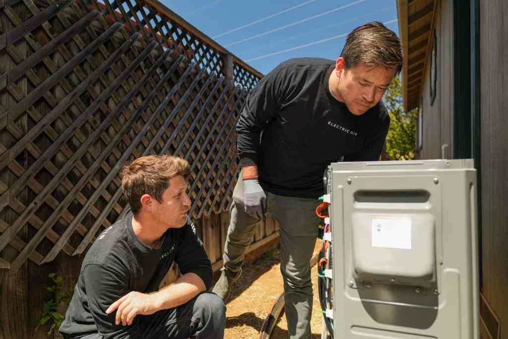 Two HVAC contractors inspecting the wiring inside an outdoor heat pump unit beside a California home.